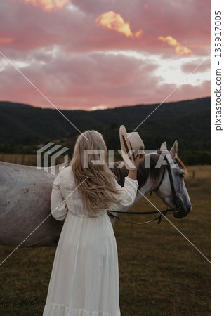Woman holding hat by gray horse during sunset on a field with clouds in the sky Woman holding hat by gray horse during sunset on a field with clouds in the sky 135917005