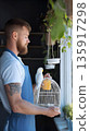Caucasian male pet shop seller gently placing a parrot back into cage . Tropical birds chirping around him. Sunlight streams in through front windows. 135917298