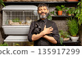 Mediterranean male pet shop seller in apron is holding black rabbit in his arms in cozy pet shop. Small aviaries with rabbits, various potted plants. 135917299