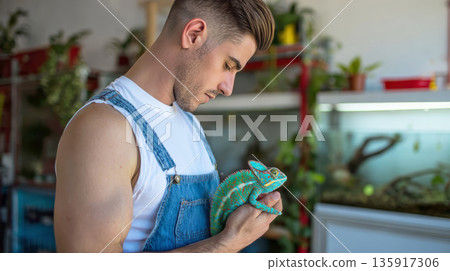 Caucasian male pet shop seller is holding a chameleon in his arms, around reptile enclosures, terrariums and exotic plants. Minimalistic background. 135917306