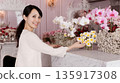 Cute smiling east asian female seller organizing pastel chrysanthemums, orchids and chamomile on a marble-top counter. Serene flower boutique. 135917308