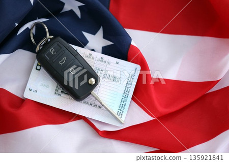 Ignition keys with remote control buttons and driver license card on United States flag. Concept of car driving in USA. Vehicle ownership Ignition keys with remote control buttons and driver license card on United States flag. Concept of car driving in USA. Vehicle ownership 135921841