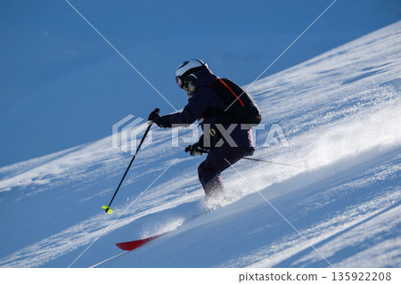 Skier in purple jacket descends snowy slope with poles and backpack on bright winter day 135922208