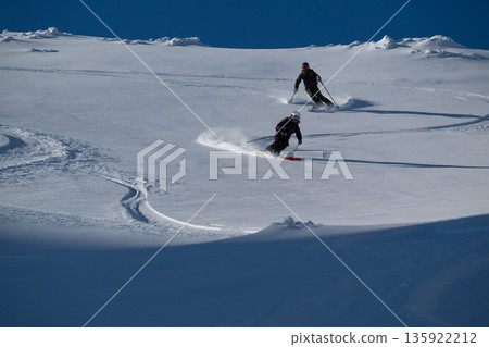 Two skiers carve a fresh off-piste descent in the pristine snowy mountains, enjoying a freeride heliskiing adventure 135922212