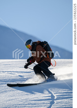 Skier in brown jacket carving a turn on a snowy slope with clear blue sky 135922225
