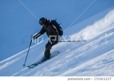 Skier descends a snowy slope with helmet and backpack on a clear blue sky day 135922227