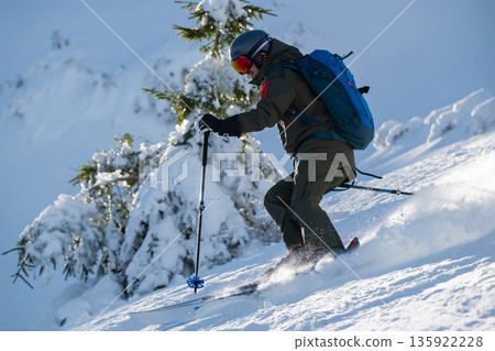 Skier in rugged backcountry snow descending a steep slope with poles and a blue backpack 135922228