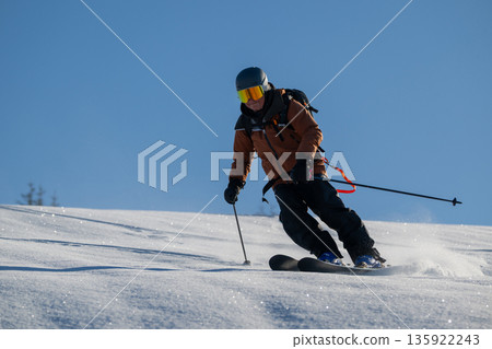 Skier in orange jacket carving down a snowy slope during off-piste freeride action under clear blue sky Skier in orange jacket carving down a snowy slope during off-piste freeride action under clear blue sky 135922243