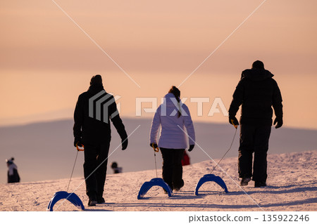 Three people walk on snowy hill at sunset pulling blue sleds, enjoying winter outdoor adventure Three people walk on snowy hill at sunset pulling blue sleds, enjoying winter outdoor adventure 135922246
