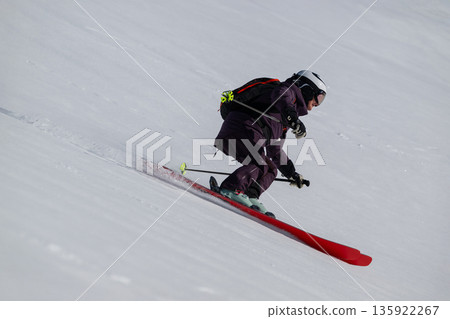 Skier in purple gear carving down a snowy slope with helmet and backpack on a bright day 135922267