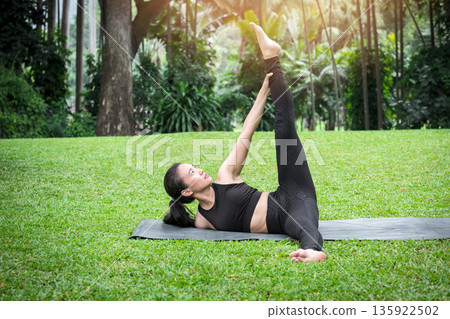 Asian woman doing side lying yoga poses to stretching leg and shoulder into a triangular shape on mat in outdoor park. Asian woman doing side lying yoga poses to stretching leg and shoulder into a triangular shape on mat in outdoor park. 135922502