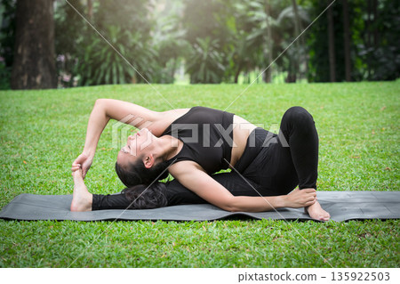 Asian woman practicing yoga in Revolved Head-to-Knee Pose on the mat in outdoor park. Asian woman practicing yoga in Revolved Head-to-Knee Pose on the mat in outdoor park. 135922503