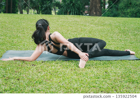 Asian woman doing side lying yoga poses to stretching leg and flank on mat in outdoor park. rear view. Asian woman doing side lying yoga poses to stretching leg and flank on mat in outdoor park. rear view. 135922504