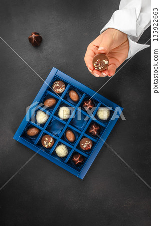 Female hand holding chocolate praline above luxury gift box with assorted chocolates Female hand holding chocolate praline above luxury gift box with assorted chocolates 135922663