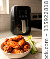 Crispy Fried Chicken Wings in a Bowl with Celery and Dip, with a Modern Black Air Fryer in the Background. Healthy Home Cooking Concept in a Bright Kitchen with Natural Sunlight. 135922818