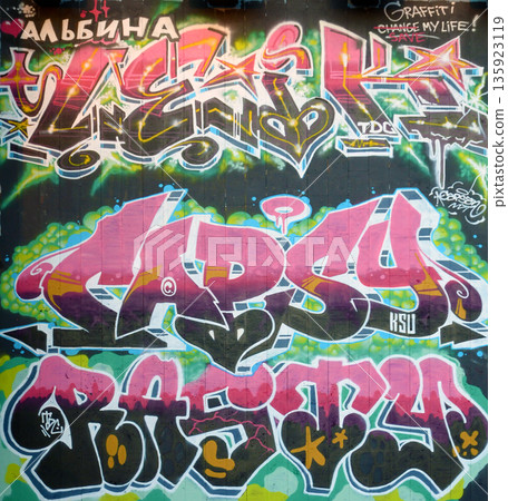 The old wall decorated with paint stains in the style of street art culture. Colorful background of full graffiti painting artwork with bright aerosol outlines on wall. Colored background texture The old wall decorated with paint stains in the style of street art culture. Colorful background of full graffiti painting artwork with bright aerosol outlines on wall. Colored background texture 135923119