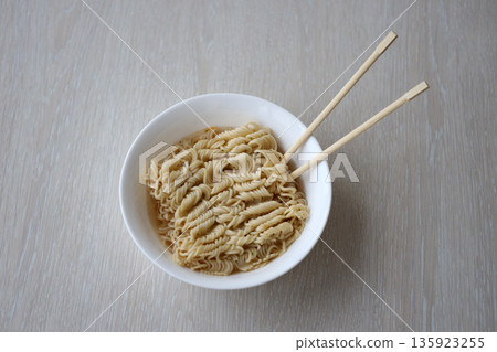 Bowl of instant cooking noodles with wooden chopsticks on white wooden table close up Bowl of instant cooking noodles with wooden chopsticks on white wooden table close up 135923255