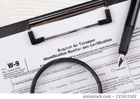 IRS Form W-9 Request for taxpayer identification number and certification, blank on A4 tablet lies on office table with pen and magnifying glass close up 135923382