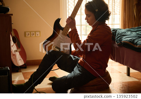 Teen Boy Plays Electric Guitar in Cozy Bedroom with Headphones On, Focused on Practice Teen Boy Plays Electric Guitar in Cozy Bedroom with Headphones On, Focused on Practice 135923502