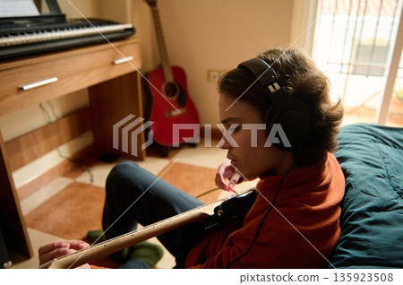 Teenage Boy Wearing Headphones Playing Guitar in Cozy Home Music Room with Keyboard Teenage Boy Wearing Headphones Playing Guitar in Cozy Home Music Room with Keyboard 135923508