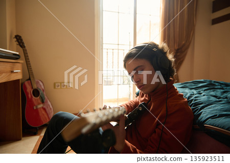Teenage Boy Playing Guitar in Bedroom with Headphones, Focused on Music Practice 135923511