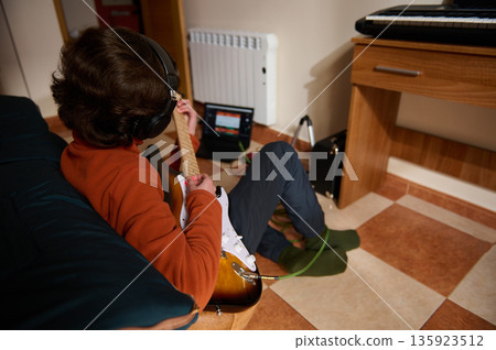 Teenage Boy Playing Electric Guitar With Headphones In Cozy Home Music Setup Teenage Boy Playing Electric Guitar With Headphones In Cozy Home Music Setup 135923512