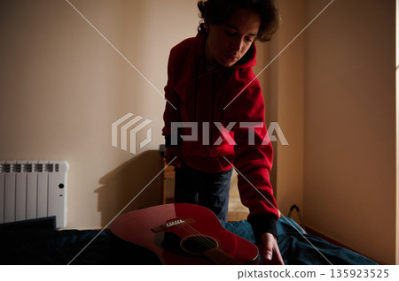 Teen Boy in Red Hoodie Adjusts Acoustic Guitar in Dim Room, Quiet Musical Moment 135923525