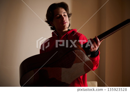 Teenager Boy In Red Hoodie Playing Acoustic Guitar In Softly Lit Indoor Setting 135923528