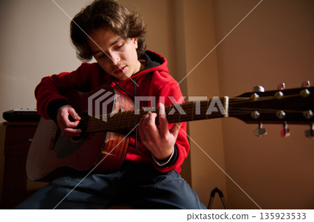 Teen Guitar Player In Red Hoodie Strums Acoustic Guitar In Quiet Indoor Setting 135923533