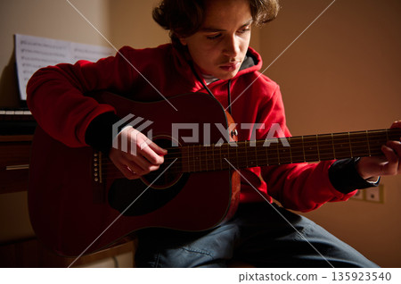 Teen Guitar Player In Red Hoodie Focused On Acoustic Guitar In Cozy Indoor Setting Teen Guitar Player In Red Hoodie Focused On Acoustic Guitar In Cozy Indoor Setting 135923540