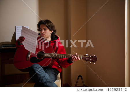 Teen Guitar Player Reads Sheet Music While Strumming Red Acoustic Guitar at Home 135923542