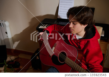 Teen Guitar Player Practicing At Home With Sheet Music And Keyboard In Backdrop Teen Guitar Player Practicing At Home With Sheet Music And Keyboard In Backdrop 135923543