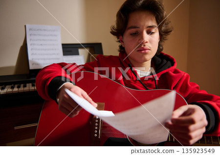 Teen Guitar Player Focused On Sheet Music With Piano In Background 135923549