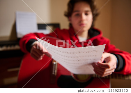 Teenage Musician Reads Sheet Music While Strumming Guitar in Cozy Home Setting Teenage Musician Reads Sheet Music While Strumming Guitar in Cozy Home Setting 135923550