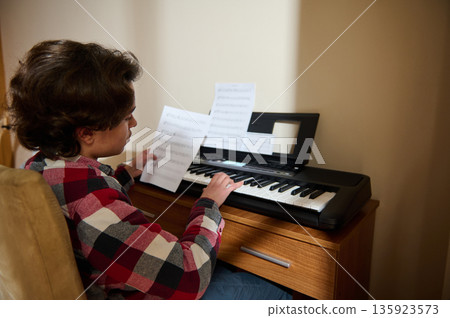 Teenager Boy Playing Piano At Home With Sheet Music On Digital Keyboard 135923573