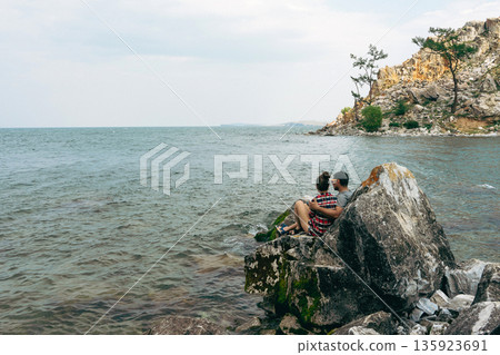 Couple, man and woman siting on rocks overlooking Baikal lake Couple, man and woman siting on rocks overlooking Baikal lake 135923691