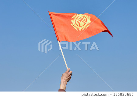 Man waving national flag from Kyrgyzstan against a clear blue sky. Patriotism and national pride concept for country celebration. Man waving national flag from Kyrgyzstan against a clear blue sky. Patriotism and national pride concept for country celebration. 135923695