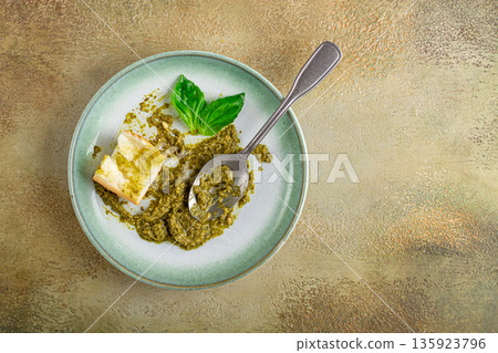 There is fresh basil pesto sauce on a platter, a slice of bread and a spoon on a plate. There is fresh basil pesto sauce on a platter, a slice of bread and a spoon on a plate. 135923796