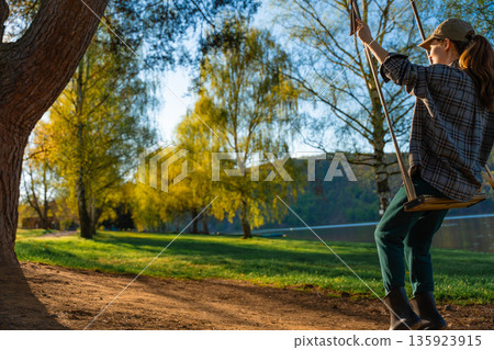 Woman in casual clothes and cap enjoying a wooden swing hung from a tree in a Czech countryside. 135923915
