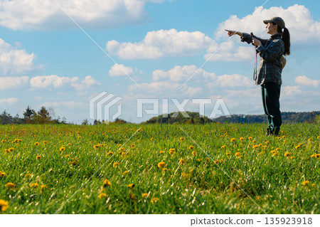 Woman holding smartphone and pointing in field of yellow dandelions Woman holding smartphone and pointing in field of yellow dandelions 135923918