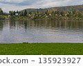 Riverside village with forested hills and cottages reflected in calm water near Nova Zivohost. 135923927
