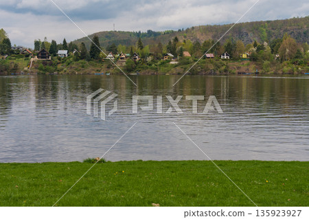 Riverside village with forested hills and cottages reflected in calm water near Nova Zivohost. 135923927