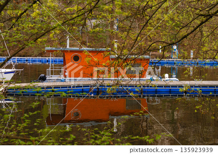 Floating house with round window docked at marina - reflection in water through spring branches 135923939