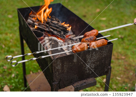 Grilling sausages over open fire on metal barbecue grill in backyard with green grass in autumn 135923947