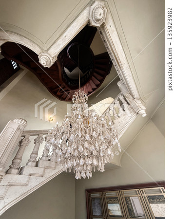 A grand chandelier hangs from the ceiling of the Baron Empain Palace in Cairo, Egypt. A spiral staircase with wooden steps is visible in the background. 135923982