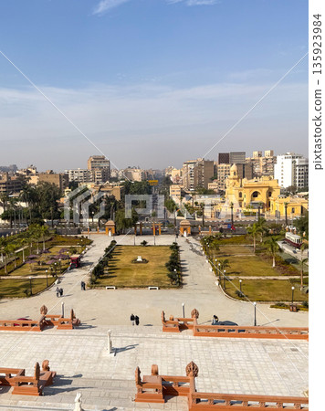 Aerial view of Heliopolis district in Cairo. The scene includes green parks, modern buildings, and clear blue skies. Urban landscape with palm trees and pathways. Aerial view of Heliopolis district in Cairo. The scene includes green parks, modern buildings, and clear blue skies. Urban landscape with palm trees and pathways. 135923984