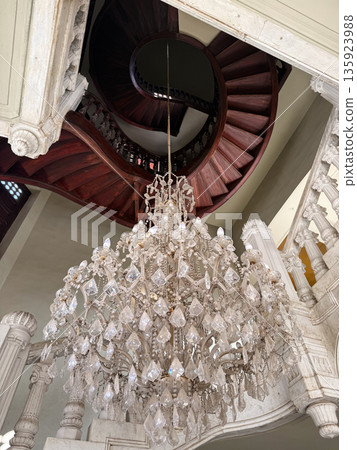 A grand chandelier hangs from the ceiling of the Baron Empain Palace in Cairo, Egypt. A spiral staircase with wooden steps is visible in the background. 135923988