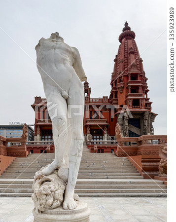 A marble statue of a headless figure stands in front of the Baron Empain Palace, an ornate red building in Cairo, Egypt. The scene captures historical architecture and art. A marble statue of a headless figure stands in front of the Baron Empain Palace, an ornate red building in Cairo, Egypt. The scene captures historical architecture and art. 135923989