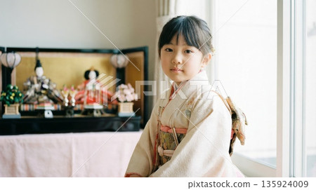 一位身著和服的美麗日本女孩站在櫥窗裡展示的精美雛人偶前,看著鏡頭。 一位身著和服的美麗日本女孩站在櫥窗裡展示的精美雛人偶前,看著鏡頭。 135924009