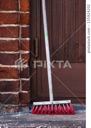 Outdoor close up of a push broom with red bristles leaning on a concrete step next to a brick wall Outdoor close up of a push broom with red bristles leaning on a concrete step next to a brick wall 135924202